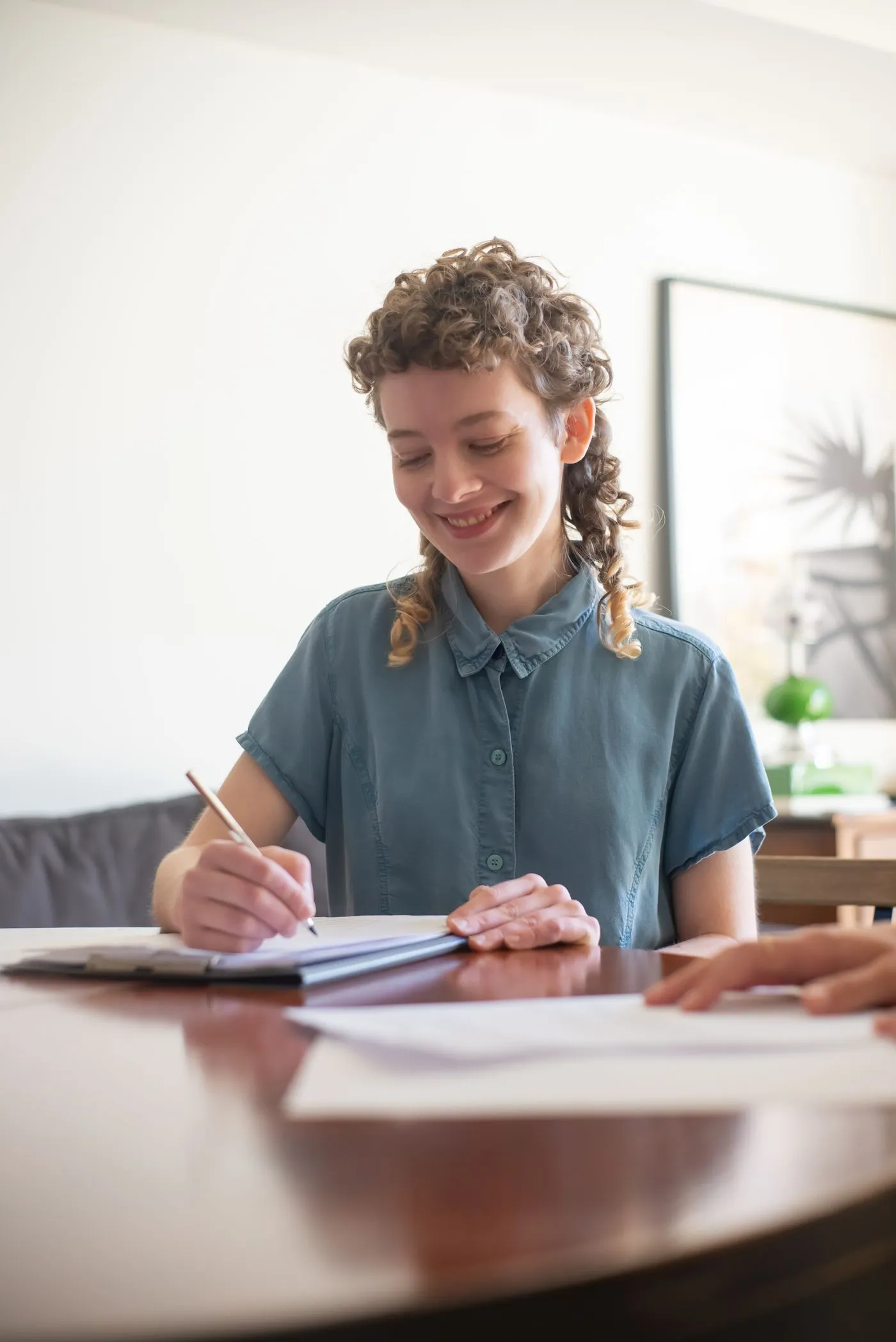 A smiling young learner writing on a clipboard at a table in a bright living room
